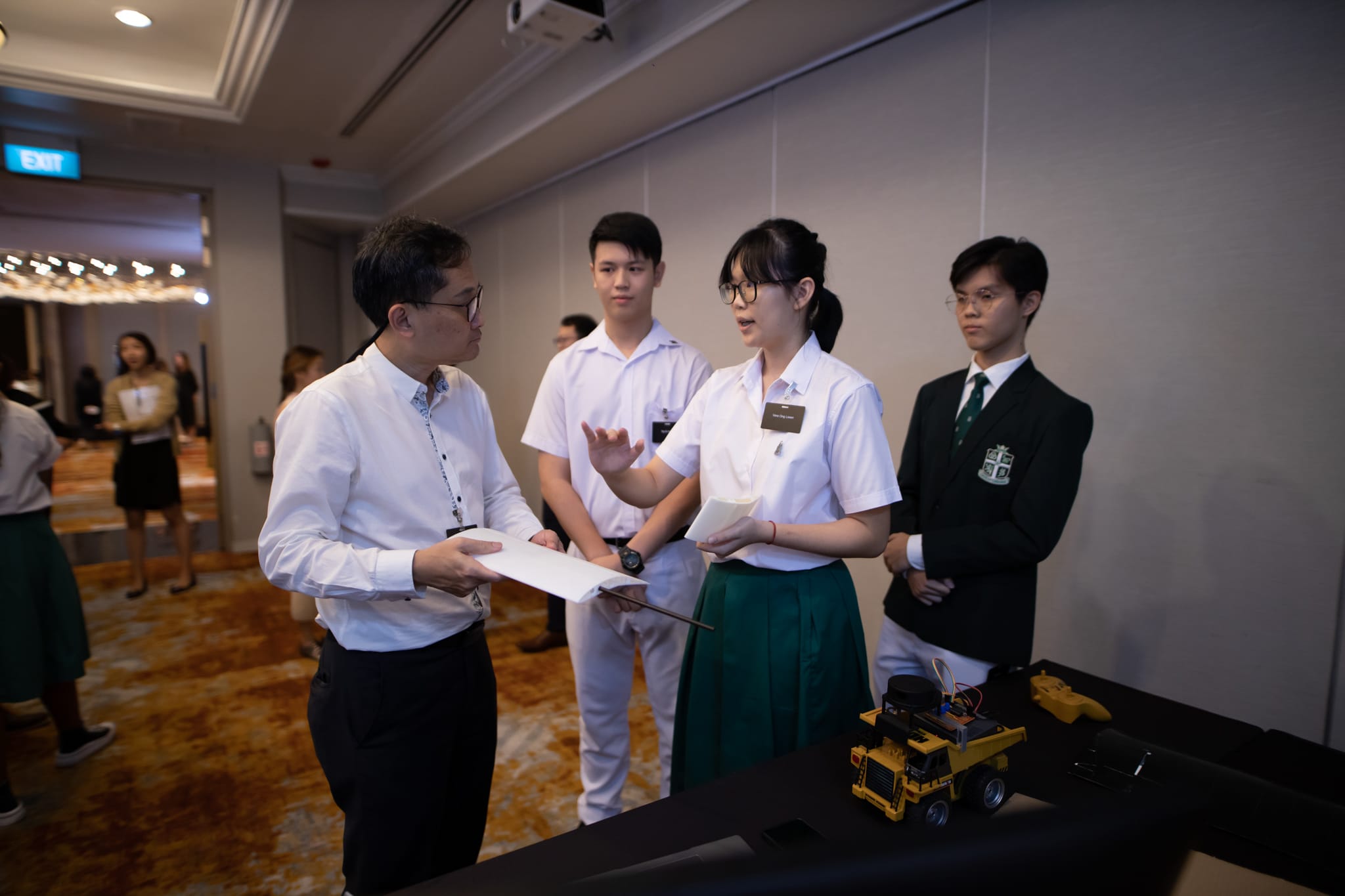 Three students in school uniforms present a toy truck project to an adult holding papers at an indoor event.